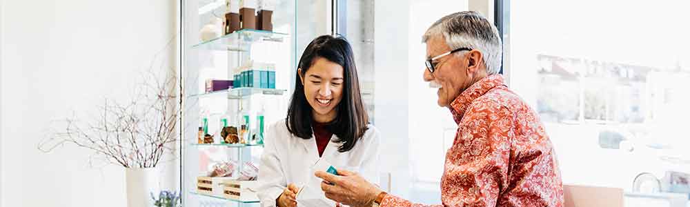 An older gray-haired white man with moustache and glasses being given a prescription by a young female pharmacist